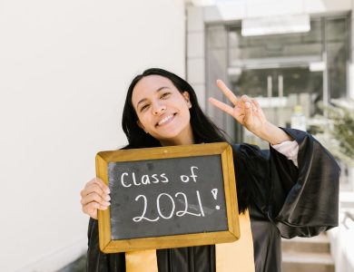 Smiling graduate in cap and gown holding a 'Class of 2021' sign while making a peace sign.