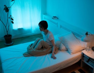Woman sitting on bed in dimly lit room, conveying themes of insomnia and contemplation.