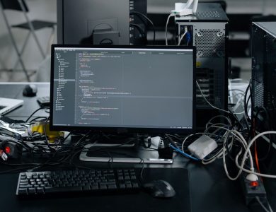 A cluttered workstation in an office featuring a monitor displaying code, surrounded by a keyboard, mouse, and wiring.