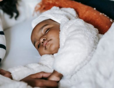 From above of crop anonymous African American mother holding hand of cute newborn baby in warm clothes sleeping on cozy bed