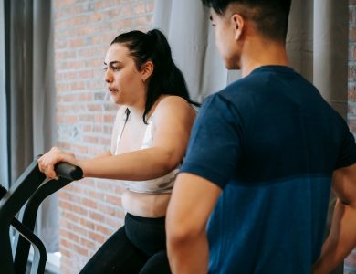Side view of serious concentrated young obese female doing weight lose exercise on cycling equipment during cardio training with crop ethnic male personal coach