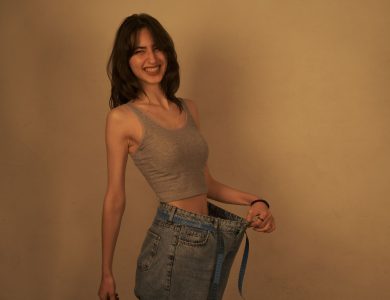 Smiling woman showcasing weight loss by wearing oversized jeans indoors.