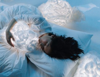 Woman in white hugging glowing cloud pillows in serene, dreamlike setting.